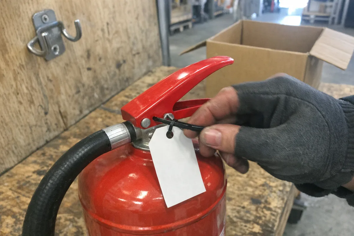 Gloved hands tightening a nylon cable tie to attach a yellow service tag through the handle of a red portable extinguisher on a packing bench in Stockholm.